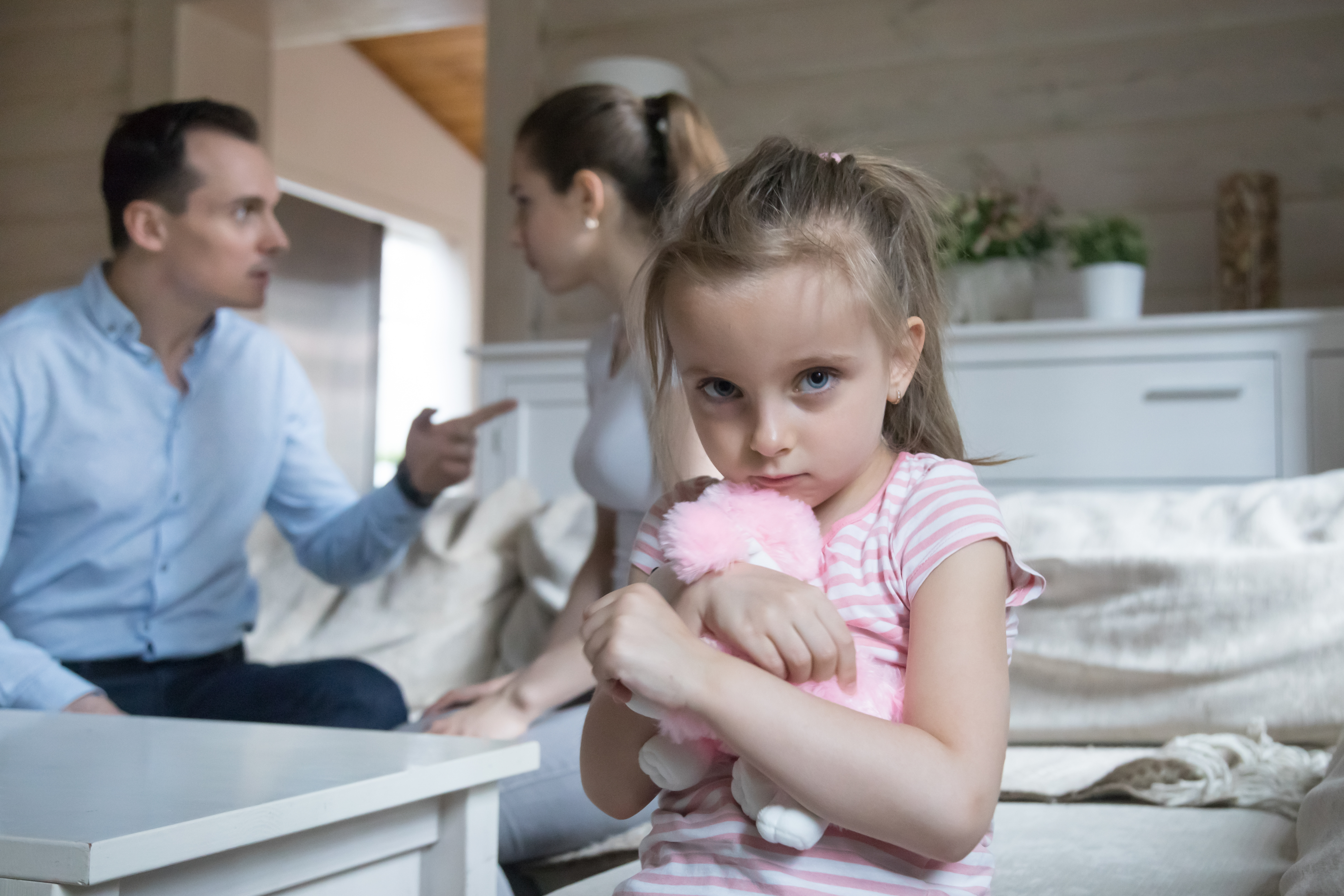 Little girl holding teddy bear while parents fight in background